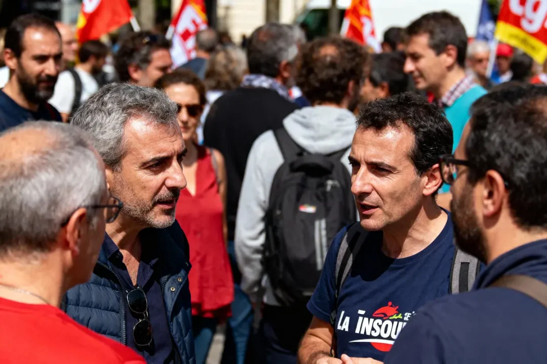 Personnes discutant lors d'une manifestation avec drapeaux en arrière-plan, ambiance animée et ensoleillée.