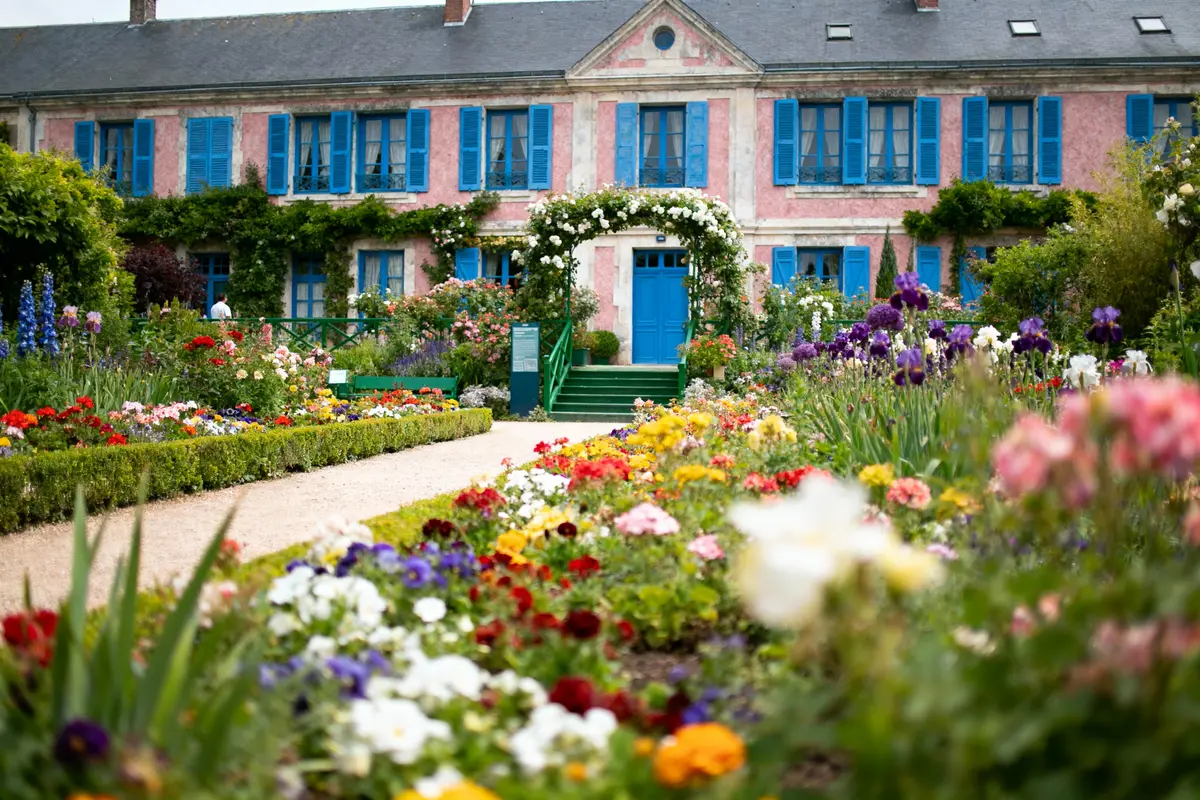 Jardin fleuri coloré devant maison rose aux volets bleus, avec allée centrale et verdure abondante.