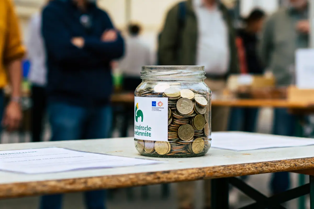 Un bocal rempli de pièces sur une table lors d'un événement, flou de personnes en arrière-plan.