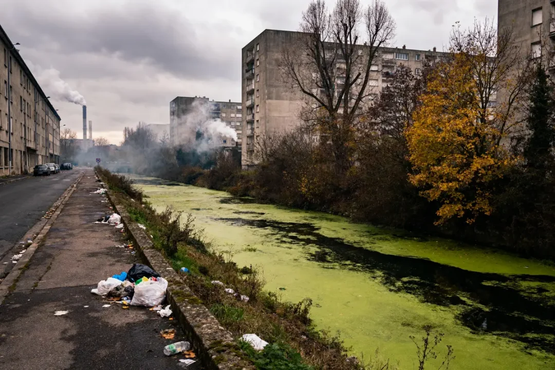 Rue urbaine avec déchets, canal pollué, bâtiments en arrière-plan, ciel nuageux, arbres aux feuilles jaunissantes.