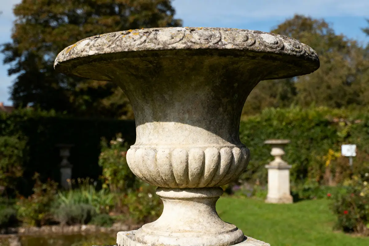 Vase en pierre ornée, dans un jardin verdoyant, avec d'autres sculptures en arrière-plan.