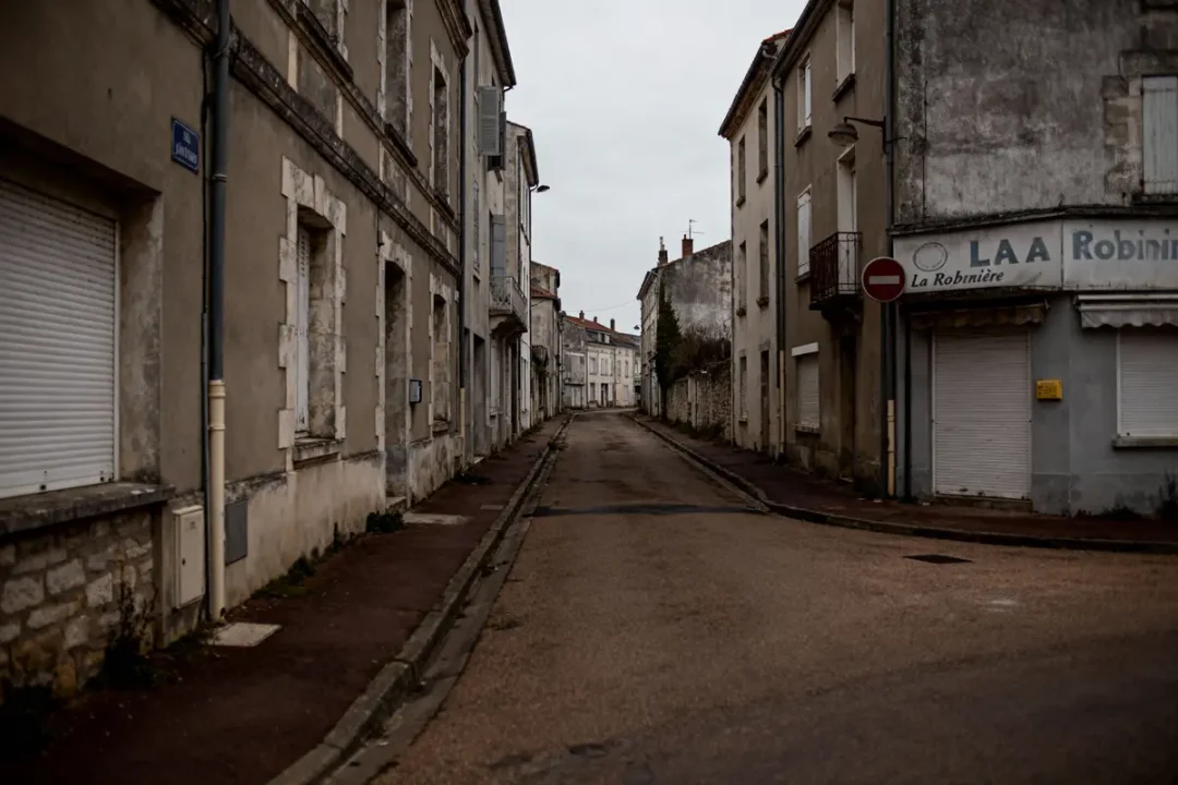 Rue déserte dans un village, bâtiments anciens, volets fermés, ciel nuageux, ambiance calme et silencieuse.