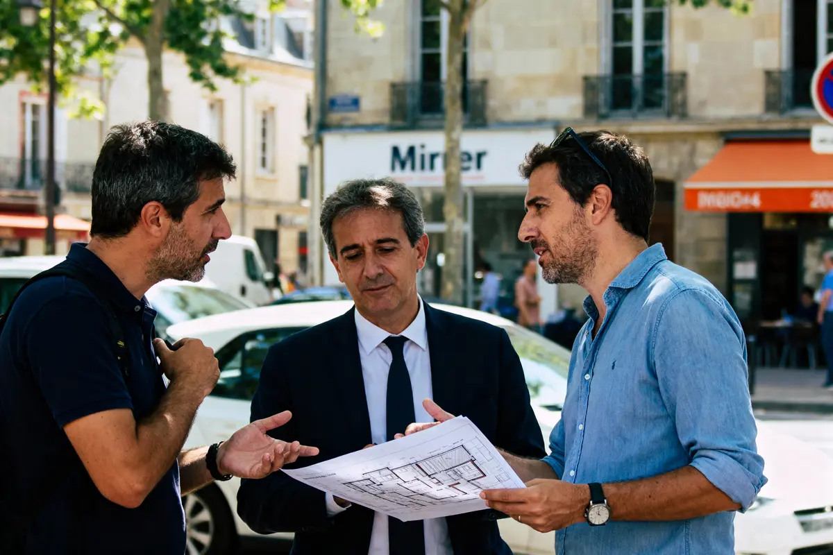 Trois hommes discutent de plans architecturaux devant un bâtiment ensoleillé, en tenue décontractée et formelle.