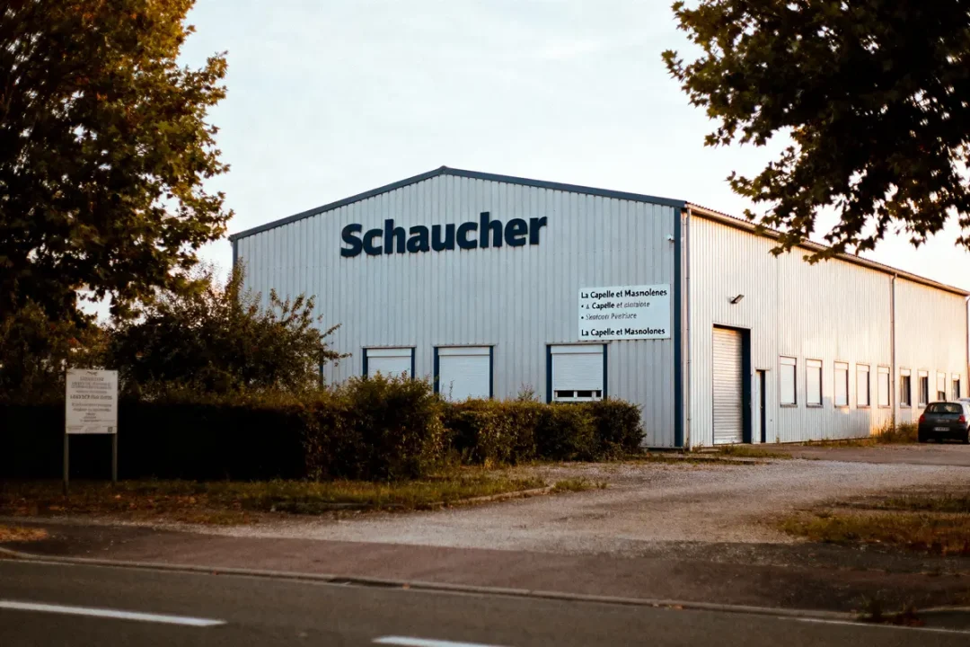Bâtiment industriel avec inscription "Schaucher", entouré de verdure, ciel clair, panneau proche, voiture stationnée à droite.