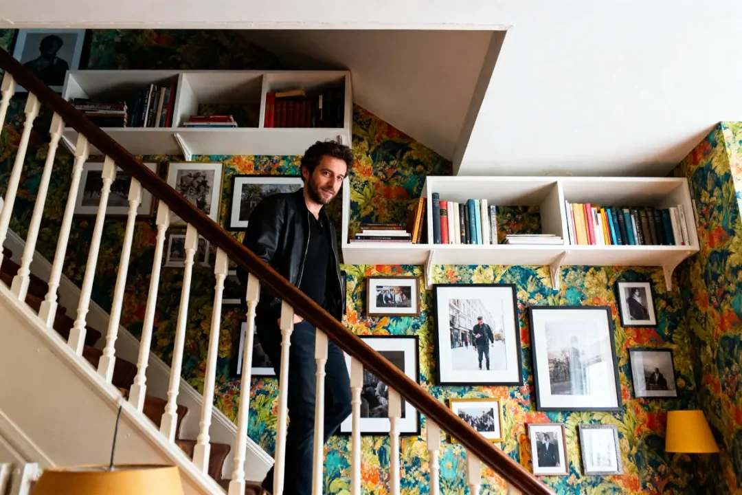 Homme sur un escalier avec mur tapissé, étagères de livres, et photos encadrées. Ambiance chaleureuse et colorée.