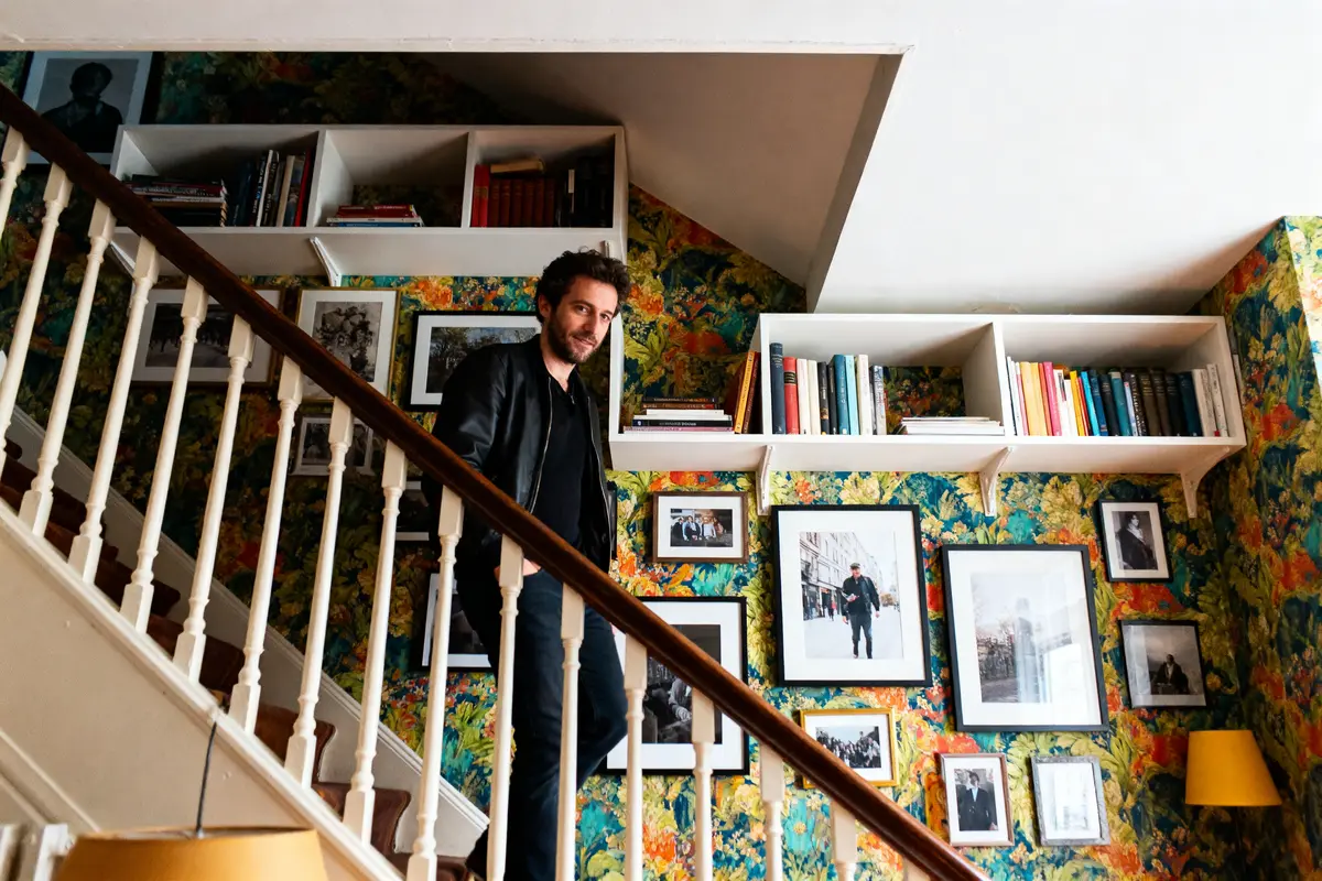 Homme sur un escalier avec mur tapissé, étagères de livres, et photos encadrées. Ambiance chaleureuse et colorée.