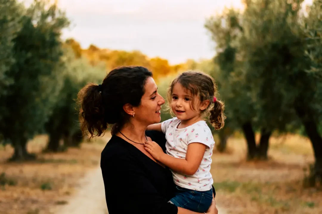 Mère souriante portant enfant dans un champ d'oliviers ensoleillé, chemin terreux en arrière-plan.