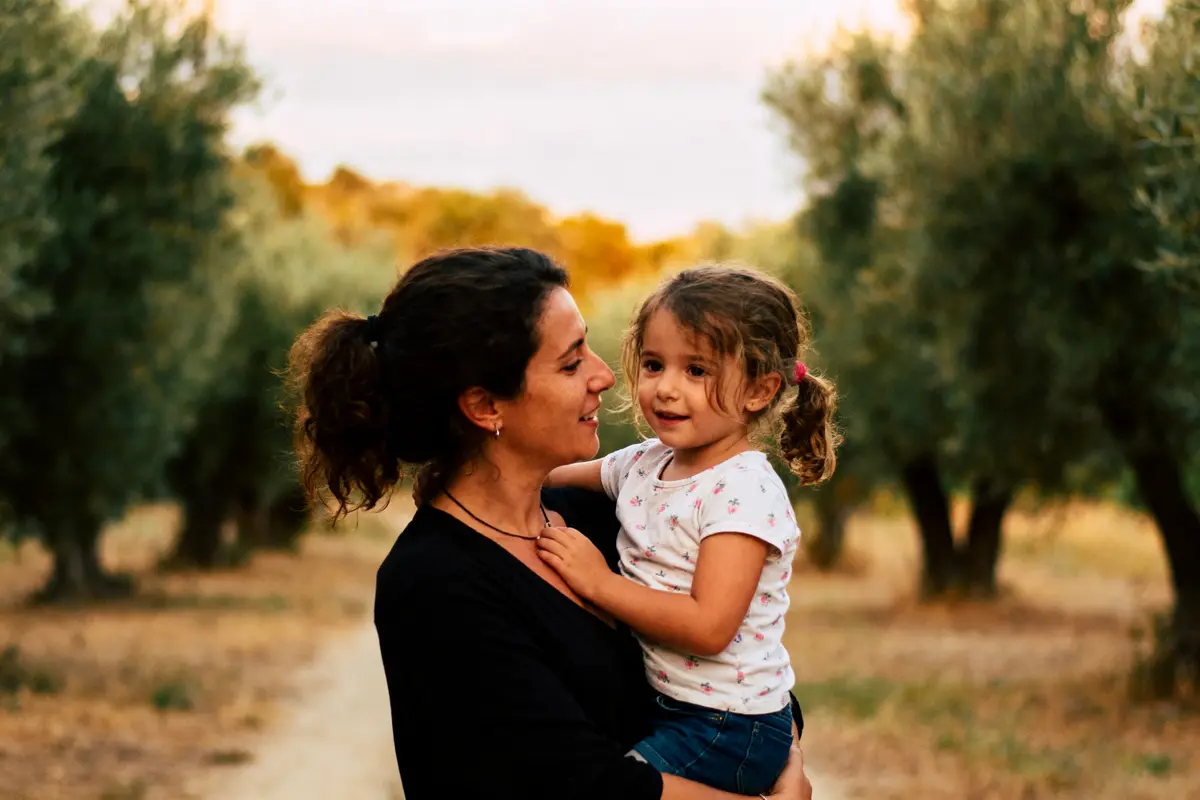 Mère souriante portant enfant dans un champ d'oliviers ensoleillé, chemin terreux en arrière-plan.