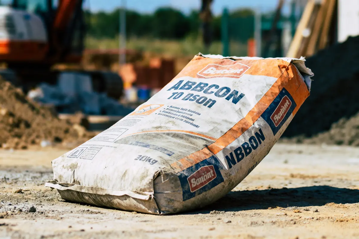 Sac de ciment "Bolarit Abbon" sur chantier, sol terreux, matériel de construction en arrière-plan flou.