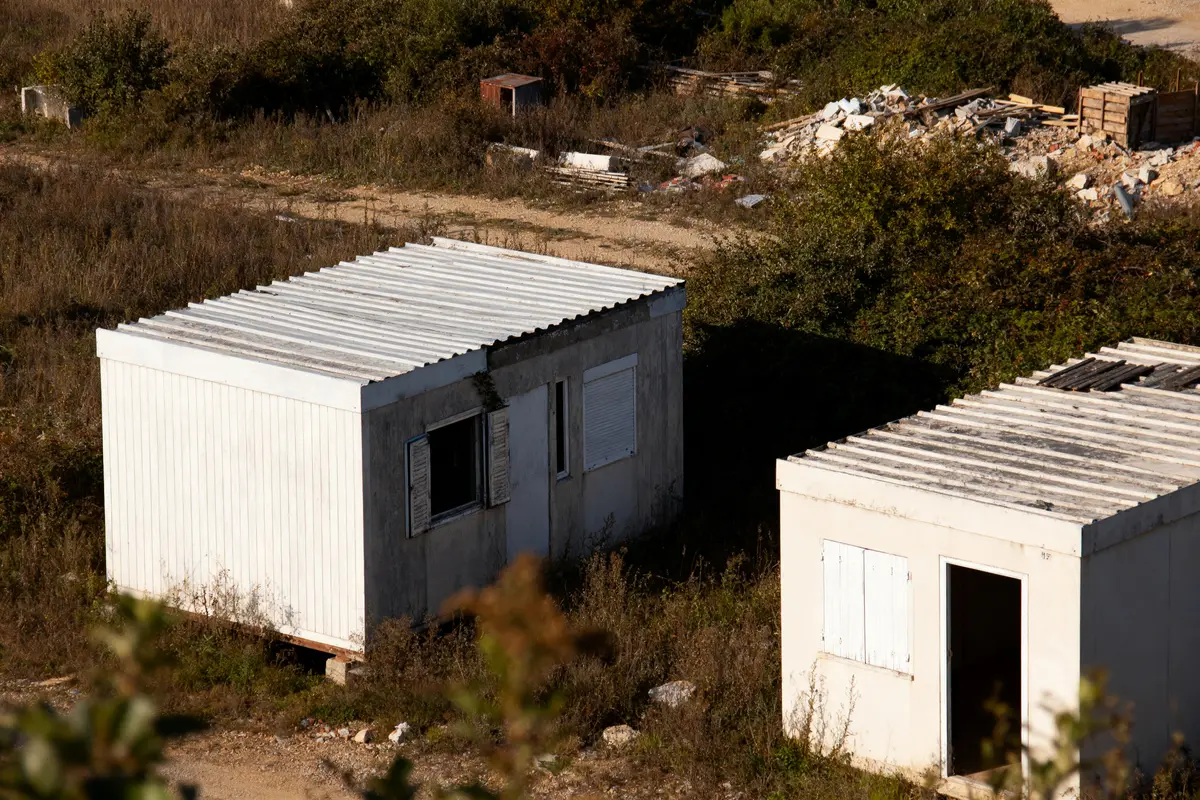 Deux cabanes blanches en métal et béton, entourées de végétation et de débris.