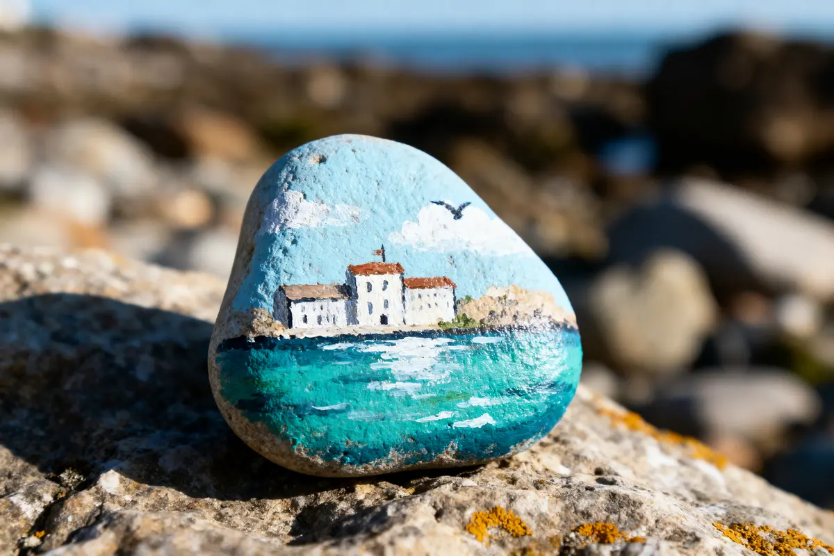 Pierre peinte représentant un paysage côtier, avec maisons blanches sous ciel bleu et mer turquoise.